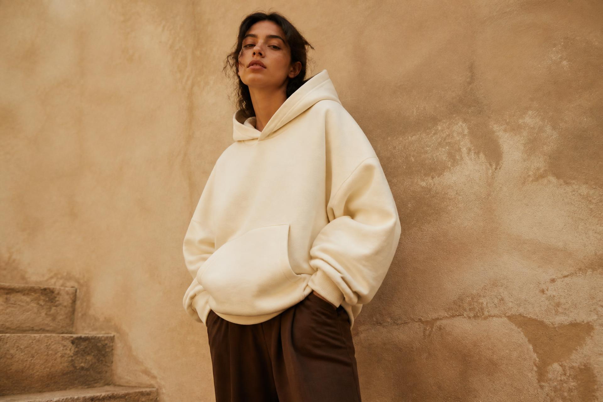 Model wearing an oversized cream hoodie against a warm plaster wall